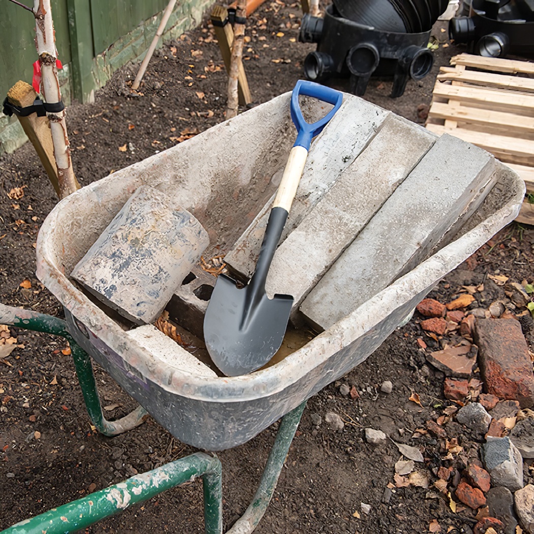 A stubby short round head shovel with a blue handle placed in a wheelbarrow with bricks and soil.