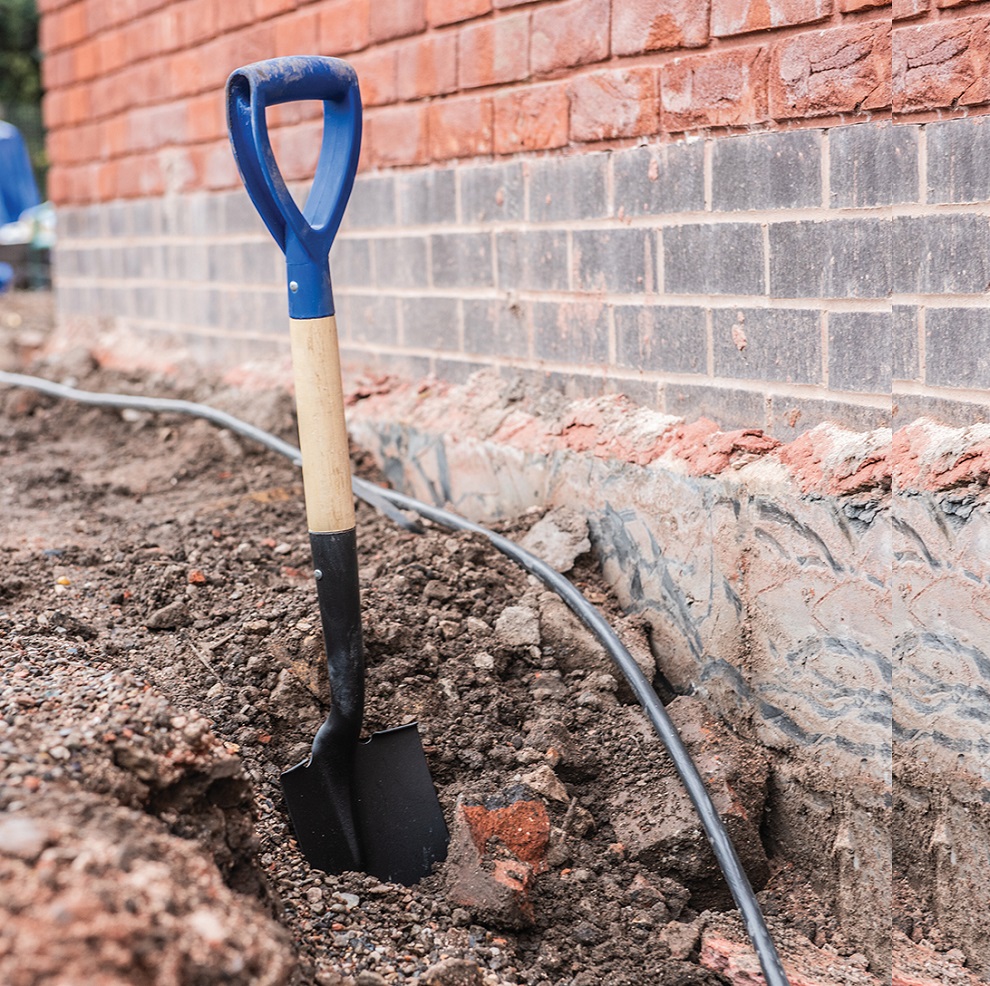 Stubby short round head shovel with blue handle in a trench by a brick wall.