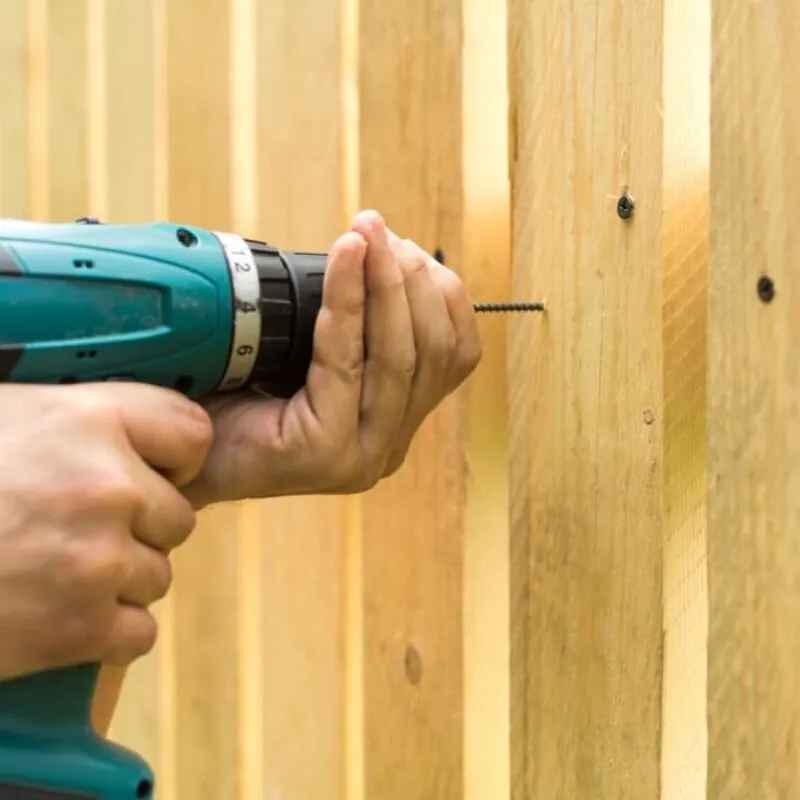 Person using a cordless drill to secure a screw into a wooden fence