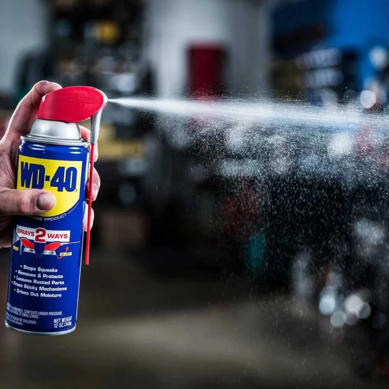 A hand holding a can of WD40 Multi-Use Maintenance Aerosol with Smart Straw, spraying in a workshop setting.