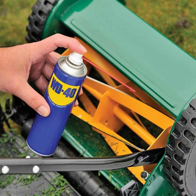 Person spraying WD-40 on a manual push mower blade to reduce grass sticking
