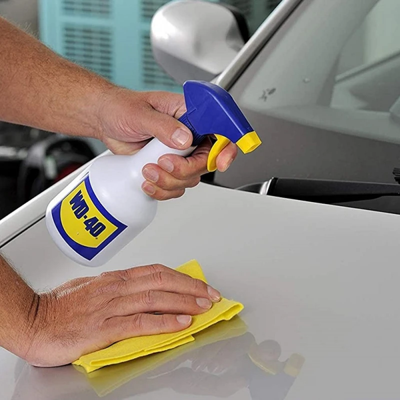 Person spraying WD-40 onto a car bonnet and wiping with a yellow cloth for surface maintenance