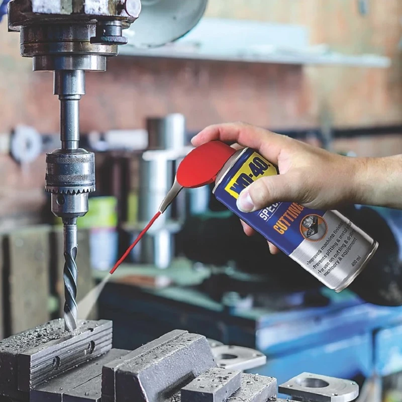 Hand using WD40 Specialist Multi-Purpose Cutting Oil Spray on a drill bit in a workshop setting.