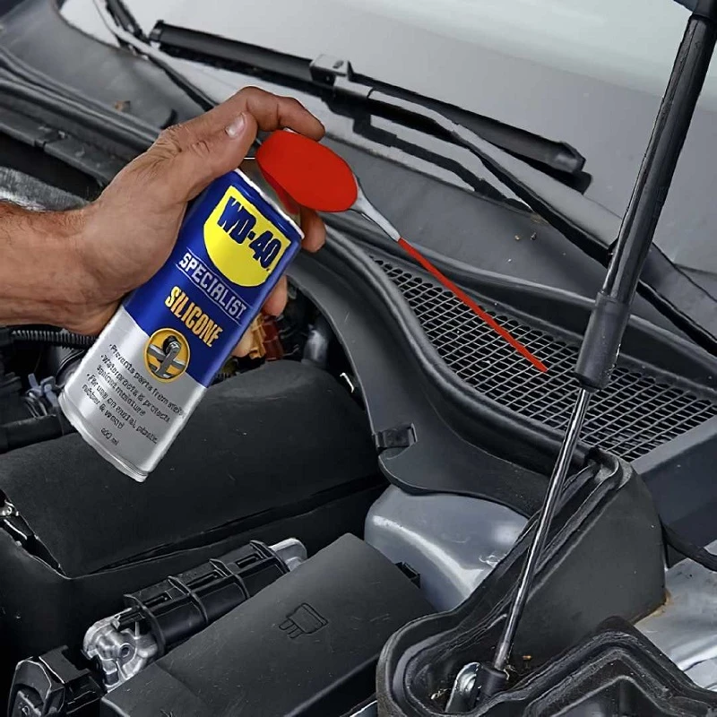 Hand using WD40 Specialist High-Performance Silicone Spray on car engine components.