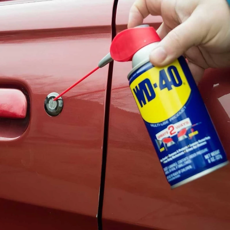 Hand using WD-40 on car door lock and rubber seals to reduce stiffness and noise