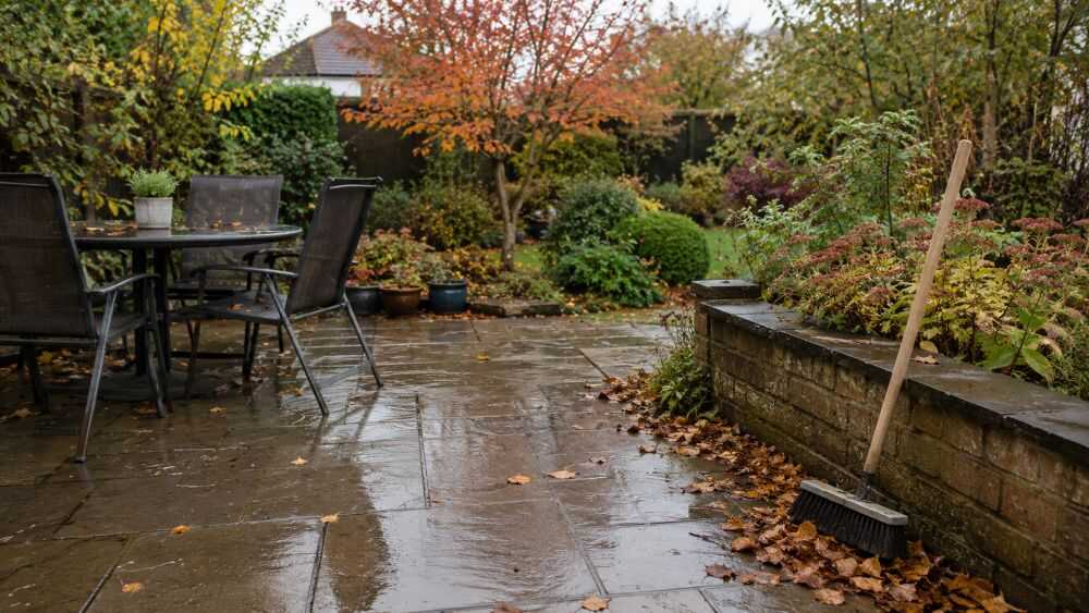 Autumn patio maintenance showing wet paving, fallen leaves near the edges and an outdoor yard brush ready for leaf clearance