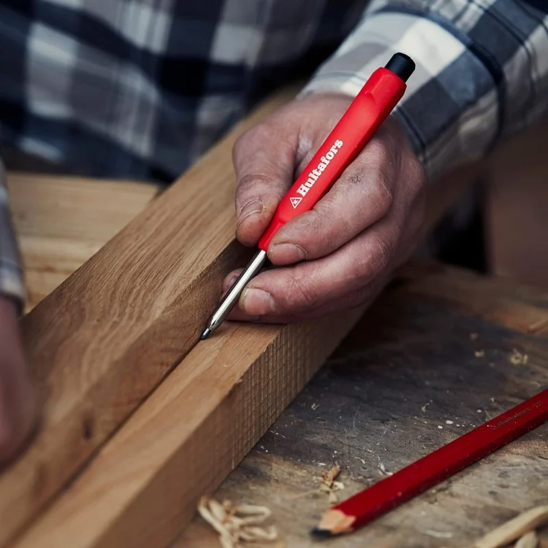 A hand using a red Hultafors dry marker to draw a line on a timber board.