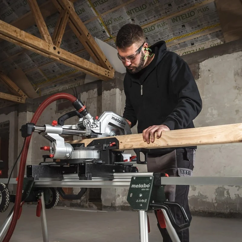 A worker using a Metabo mitre saw to cut a timber board supported on a work stand.
