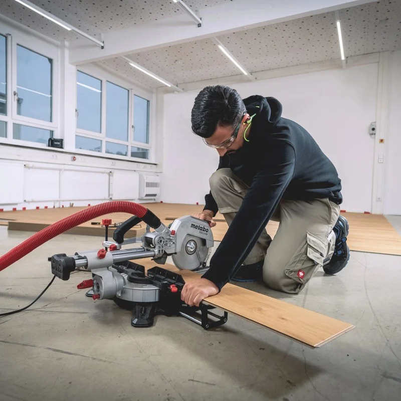 A worker operating a Metabo mitre saw at floor level with a timber board extending to the right.