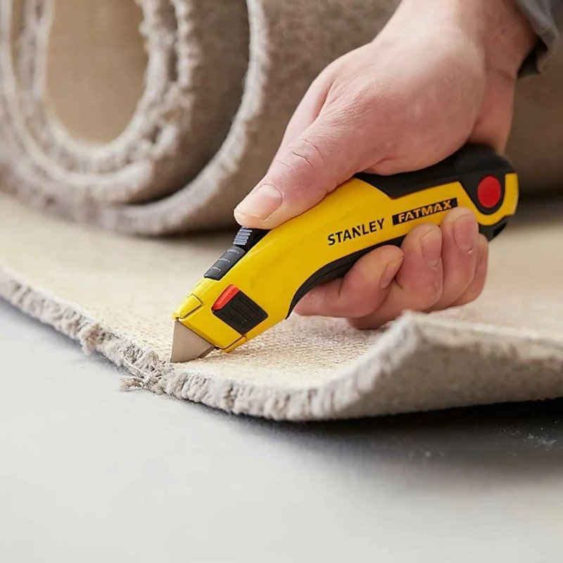 A hand using a yellow Stanley utility knife to cut carpet along its edge.